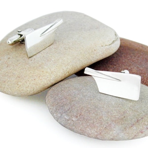 May include: A pair of silver cufflinks shaped like an oar blade. The cufflinks are sitting on two smooth, gray stones.