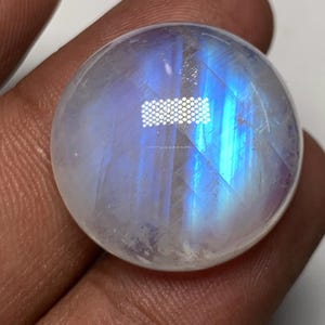 Puede incluir: Una piedra lunar redonda y pulida con una base blanca lechosa translúcida y destellos iridiscentes azules y morados. La piedra se sostiene en una mano, mostrando su superficie lisa y reflectante. La piedra preciosa es un corte cabujón.