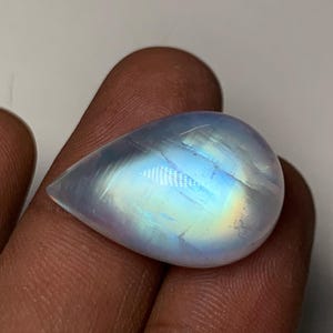 May include: A pear-shaped, polished moonstone gemstone. The stone has a translucent, milky white base with iridescent flashes of blue, yellow, and purple. The gemstone is held in a hand.