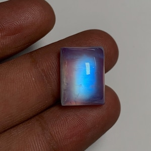 May include: A rectangular, iridescent gemstone with a polished surface. The stone displays a gradient of colors, including blue, purple, and pink. The gemstone is held in a hand, showcasing its size and shape. This item is suitable for jewelry making.