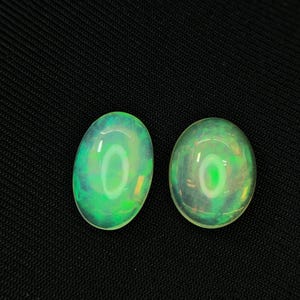 May include: Two oval-shaped cabochon gemstones with a translucent, milky white base and vibrant flashes of green and yellow play-of-colour. The stones have a smooth, polished surface that reflects light, creating a bright, circular highlight on each.