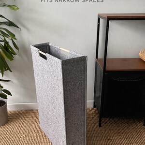 May include: A tall, rectangular gray felt laundry hamper with wooden handles. The hamper is designed to fit narrow spaces. Text on the wall reads "READY TO SHIP" and "FITS NARROW SPACES".