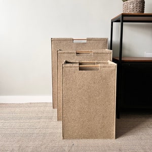 Puede incluir: Tres contenedores de almacenamiento rectangulares beige con asas de madera están alineados. Los contenedores están hechos de un material texturizado y son de diferentes tamaños. También se ven dos colgantes de pared tejidos y una suculenta en maceta.