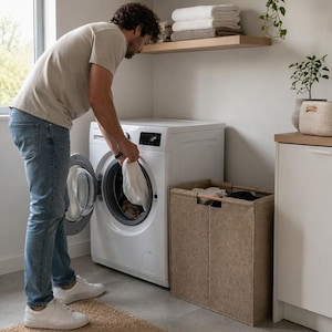 May include: A person loading a white washing machine in a laundry room. A beige woven rug is on the floor. A brown laundry hamper is next to the washing machine. Shelves with towels and plants are on the wall.