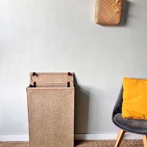 May include: A rectangular, open cardboard box with a natural brown finish stands on a woven jute rug. A gray chair with wooden legs and a yellow pillow sits to the right. A woven wall decoration hangs above.