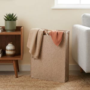 May include: A beige rectangular laundry hamper with a wooden frame and two folded blankets. The hamper is made of a textured fabric and is placed on a woven rug. A small succulent plant in a green pot sits on a wooden side table.