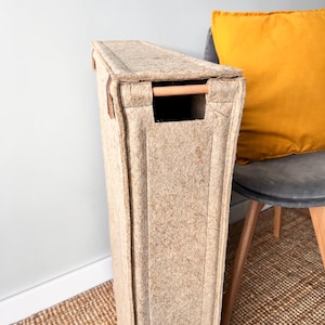 May include: A tall, rectangular storage bin made of a light brown, textured material. It features a wooden handle and leather accents. The bin is positioned next to a gray chair with a yellow pillow.