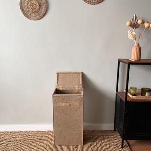 May include: A tall, rectangular, light brown storage bin with a hinged lid and a handle. The bin is made of a textured material and sits on a woven jute rug. Two round woven wall decorations are visible in the background.