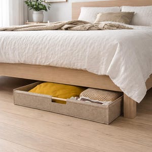 May include: A light wood bed frame with a white linen duvet cover and pillows. A beige storage drawer is pulled out from under the bed, containing a yellow pillow and a woven item. The floor is light wood.