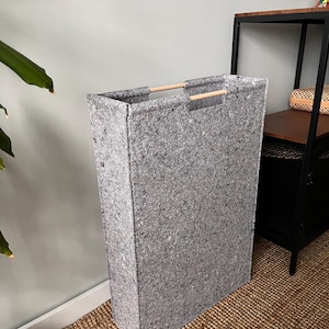 Könnte beinhalten: Ein hoher, rechteckiger, grauer Wäschekorb mit einer gesprenkelten Textur. Er hat zwei Holzgriffe oben für einfaches Tragen. Der Korb besteht aus einem robusten Material und ist so konzipiert, dass er aufrecht steht. Er steht auf einem gewebten Teppich.