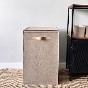 May include: A beige felt storage bin with a gold metal handle. The bin is sitting on a woven rug.