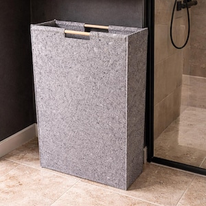 May include: A rectangular gray felt laundry hamper with wooden handles. The hamper is in a bathroom setting, next to a shower. A wooden shelf with toiletries is above the hamper.
