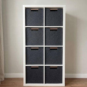 Può includere: Un mobile contenitore bianco con otto contenitori in tessuto grigio scuro. Ogni contenitore ha una maniglia in legno. L'unità è progettata per organizzare e riporre oggetti in una casa o in un ufficio. I contenitori sono uniformemente distanziati.