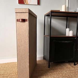 Pode incluir: Um recipiente de armazenamento alto e retangular feito de material de feltro castanho claro. O recipiente possui uma tampa a condizer e uma única pega de couro castanho fixada na lateral com costuras visíveis.