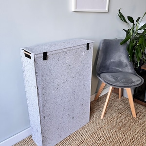 May include: A tall, grey, rectangular storage bin with a lid and black handles. The bin is made of a textured material that resembles recycled paper. The bin is sitting on a light brown rug.