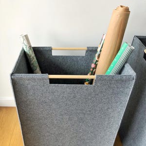 Op de afbeelding: Een grijze vilt opbergmand met een houten handvat, met rollen cadeaupapier. De mand is rechthoekig met een trapeziumvorm en staat op een houten vloer. De cadeaupapierrollen hebben verschillende kleuren en patronen.