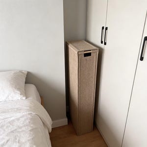 Könnte beinhalten: Eine hohe, rechteckige Aufbewahrungsbox aus beigem Stoff. Sie hat eine rechteckige Öffnung oben und einen kleinen Griffausschnitt. Die Box steht zwischen einer weißen Wand und einem weißen Schrank.