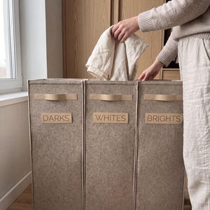May include: A beige three-section laundry hamper made of felted material. Each section features a horizontal handle and a rectangular label with the text DARKS, WHITES, and BRIGHTS printed in black. The structure is tall and rectangular.