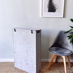 May include: A white storage bin with black straps, standing on a woven rug. The bin is made of recycled paper and has a speckled texture.