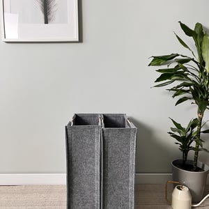 May include: Two grey felt storage bins with wooden handles. The rectangular bins stand upright on a light-coloured rug. The bins are next to a potted plant and a wall with a framed picture.