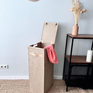 Puede incluir: Un cesto de ropa de fieltro beige con tapa, asa de cuero y una toalla rosa en su interior. El cesto está sobre una alfombra tejida frente a una pared blanca.