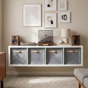May include: A contemporary living room arrangement with a white floating shelf holding grey storage bins with wooden handles. Above, framed abstract art in neutral tones adorn a beige wall. A record player, speakers, and a lamp complete the decor.