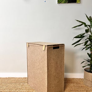Puede incluir: Un cesto de ropa de fieltro beige con asas de cuero y una lengüeta de extracción negra. El cesto está sobre una alfombra tejida.