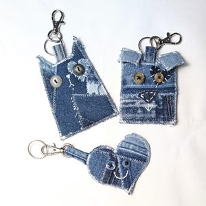 May include: Three denim keychains featuring patchwork designs. One is a cat shape, one is a square animal shape, and one is a heart shape. Each is constructed from blue denim fabric scraps with visible white stitching, button eyes, and metal clasps.