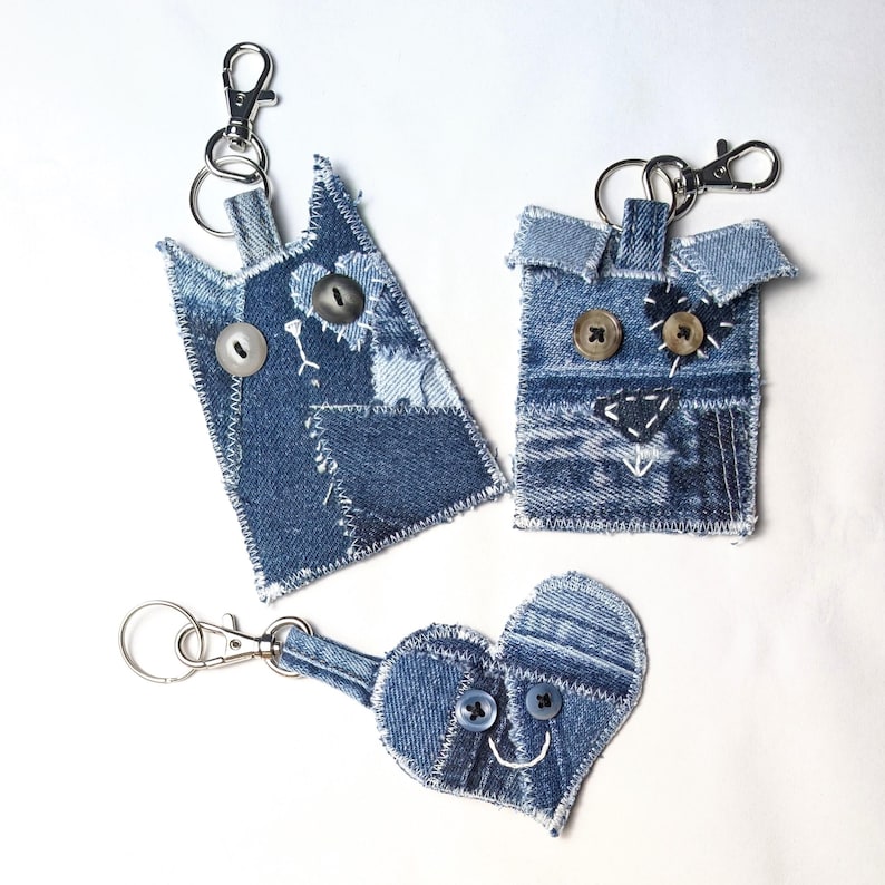 May include: Three denim keychains featuring patchwork designs. One is a cat shape, one is a square animal shape, and one is a heart shape. Each is constructed from blue denim fabric scraps with visible white stitching, button eyes, and metal clasps.