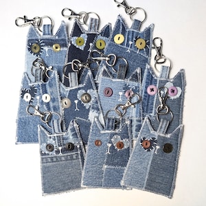 May include: A collection of cat-shaped keychains crafted from various shades of blue denim fabric. Each keychain features button eyes, stitched details, and a silver-toned metal lobster clasp attached to the top for secure hanging and utility.