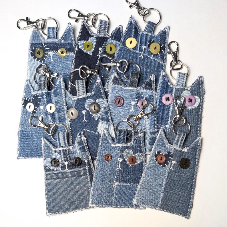May include: A collection of cat-shaped keychains crafted from various shades of blue denim fabric. Each keychain features button eyes, stitched details, and a silver-toned metal lobster clasp attached to the top for secure hanging and utility.