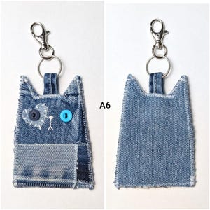 May include: A denim cat-shaped keychain labeled A6, featuring a dark blue button eye, a light blue button eye, and a white stitched heart patch. The keychain includes a silver metal lobster clasp and a split ring attached to the top loop.