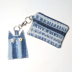 May include: A blue denim coin purse with a zipper closure attached to a cat-shaped denim keychain. The cat keychain features two yellow button eyes and stitched details, connected to the purse by a silver-toned metal split ring and lobster clasp.