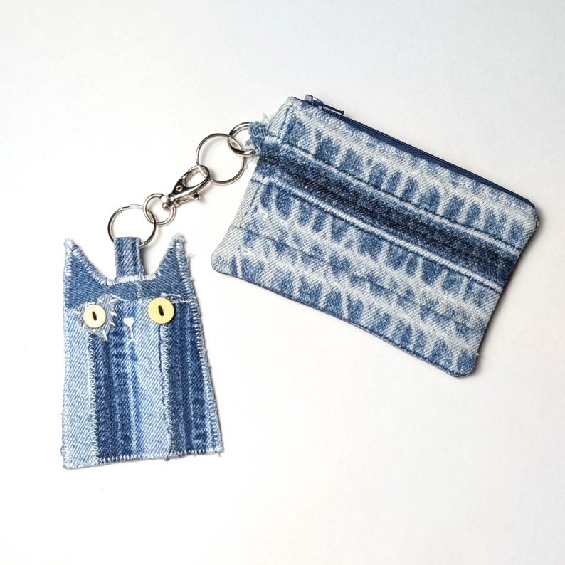 May include: A blue denim coin purse with a zipper closure attached to a cat-shaped denim keychain. The cat keychain features two yellow button eyes and stitched details, connected to the purse by a silver-toned metal split ring and lobster clasp.
