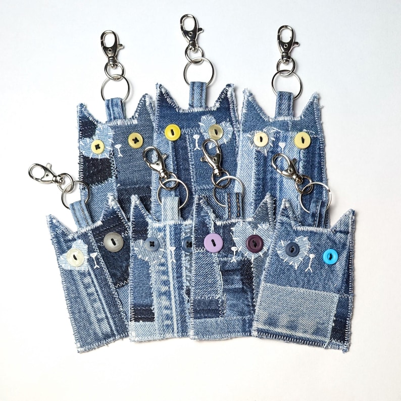 May include: A collection of seven cat-shaped keychains crafted from various shades of blue denim fabric. Each keychain features stitched white thread details, metal lobster claw clasps, and colorful plastic buttons serving as eyes.