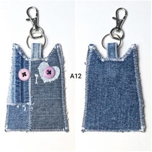 May include: A cat-shaped keychain crafted from blue denim fabric with frayed edges and white zigzag stitching. The front features two light purple buttons as eyes and a heart-shaped patch. A silver metal lobster clasp is attached. Text A12 is visible.