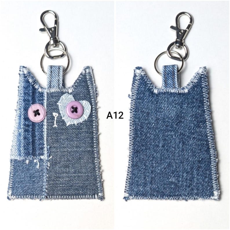 May include: A cat-shaped keychain crafted from blue denim fabric with frayed edges and white zigzag stitching. The front features two light purple buttons as eyes and a heart-shaped patch. A silver metal lobster clasp is attached. Text A12 is visible.