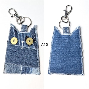 May include: A denim cat-shaped keychain featuring two yellow button eyes, white stitched details, and a silver metal lobster clasp. The front displays the text A10, while the back shows a plain blue denim surface with white zigzag stitching.