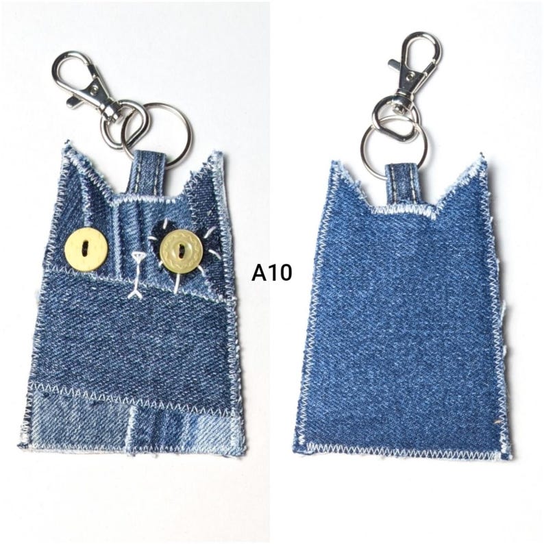 May include: A denim cat-shaped keychain featuring two yellow button eyes, white stitched details, and a silver metal lobster clasp. The front displays the text A10, while the back shows a plain blue denim surface with white zigzag stitching.