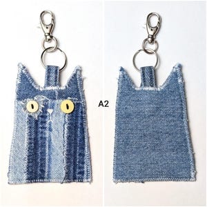 May include: A cat-shaped keychain made from blue denim fabric with frayed edges. The front features two yellow button eyes, white stitched details, and a metal lobster clasp. The back is plain blue denim. The text A2 is visible in the center.
