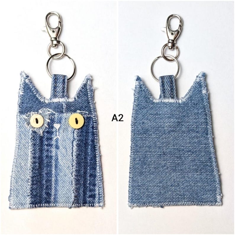 May include: A cat-shaped keychain made from blue denim fabric with frayed edges. The front features two yellow button eyes, white stitched details, and a metal lobster clasp. The back is plain blue denim. The text A2 is visible in the center.