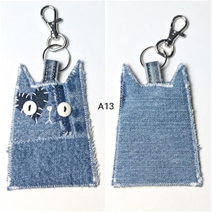 May include: A cat-shaped keychain crafted from blue denim fabric with frayed edges. The front features a dark blue denim heart patch, two white button eyes, and white stitched details. A silver metal lobster clasp is attached to the top.