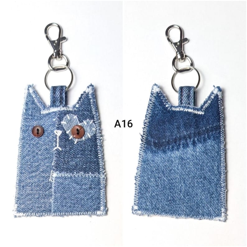 May include: A denim cat-shaped keychain labeled A16, featuring two brown button eyes, a heart-shaped patch, white stitched details, and a silver metal lobster clasp. The reverse side shows a plain blue denim fabric with white stitched edges.