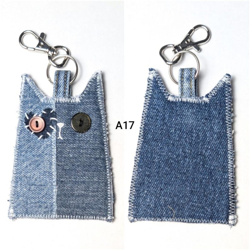 May include: A cat-shaped keychain made of blue denim fabric with a silver metal lobster clasp. The front features a light blue denim patch with a pink button eye, a dark blue denim side with a black button eye, and white stitching. Text A17 is visible.