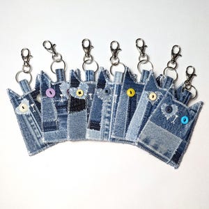 May include: A collection of eight cat-shaped keychains crafted from blue denim fabric. Each keychain features a metal lobster clasp, a split ring, and a single colorful button for an eye, with white stitched details accentuating the cat ears.