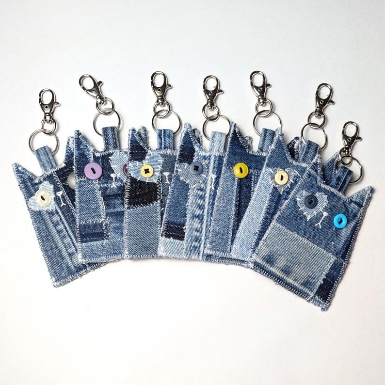 May include: A collection of eight cat-shaped keychains crafted from blue denim fabric. Each keychain features a metal lobster clasp, a split ring, and a single colorful button for an eye, with white stitched details accentuating the cat ears.