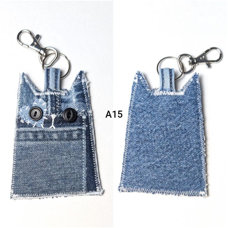 May include: A denim cat-shaped keychain with a silver metal lobster clasp. The front features two black button eyes, white stitched details, and a pocket design. The back is plain blue denim. The text A15 is visible between the two items.