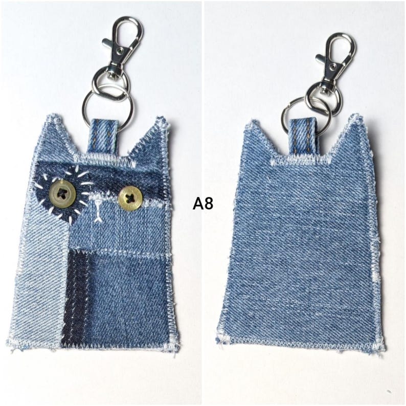 May include: A cat-shaped keychain crafted from blue denim fabric patches with white stitched edges. The front features two round buttons as eyes, one surrounded by a white stitched heart, and a silver metal lobster clasp with a split ring.