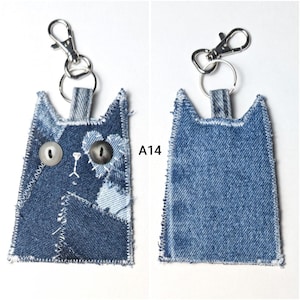 May include: A cat-shaped keychain made from patchwork denim fabric with frayed edges and white stitching. The front features two mismatched buttons for eyes and embroidered details. A silver metal lobster clasp is attached to the top. Text A14 is visible.
