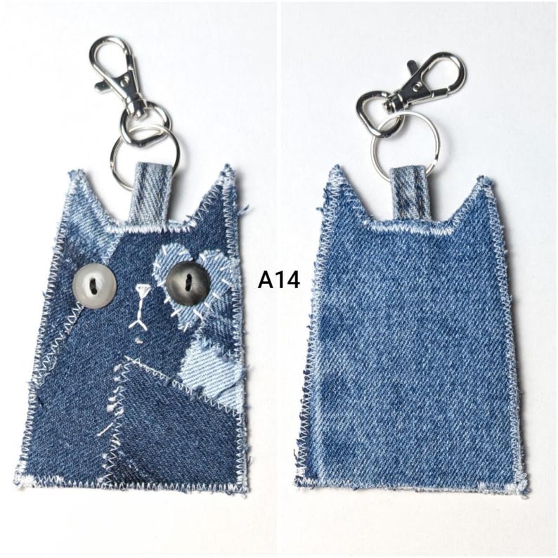 May include: A cat-shaped keychain made from patchwork denim fabric with frayed edges and white stitching. The front features two mismatched buttons for eyes and embroidered details. A silver metal lobster clasp is attached to the top. Text A14 is visible.